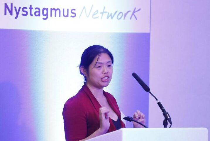 Helena Lee presents from a lectern. The Nystagmus Network logo appears on the screen behind her.