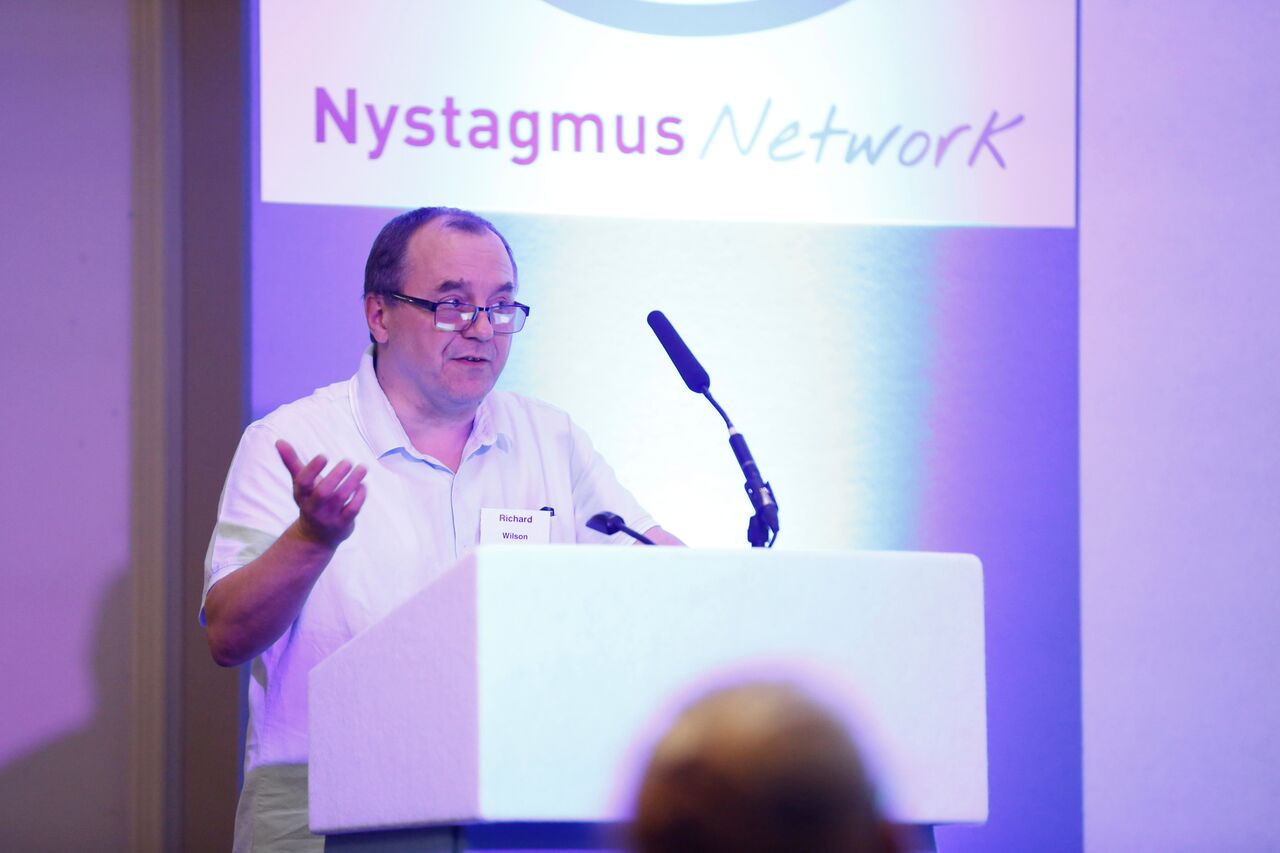Richard speaks from a podium, wearing a white Nystagmus Network T-shirt.