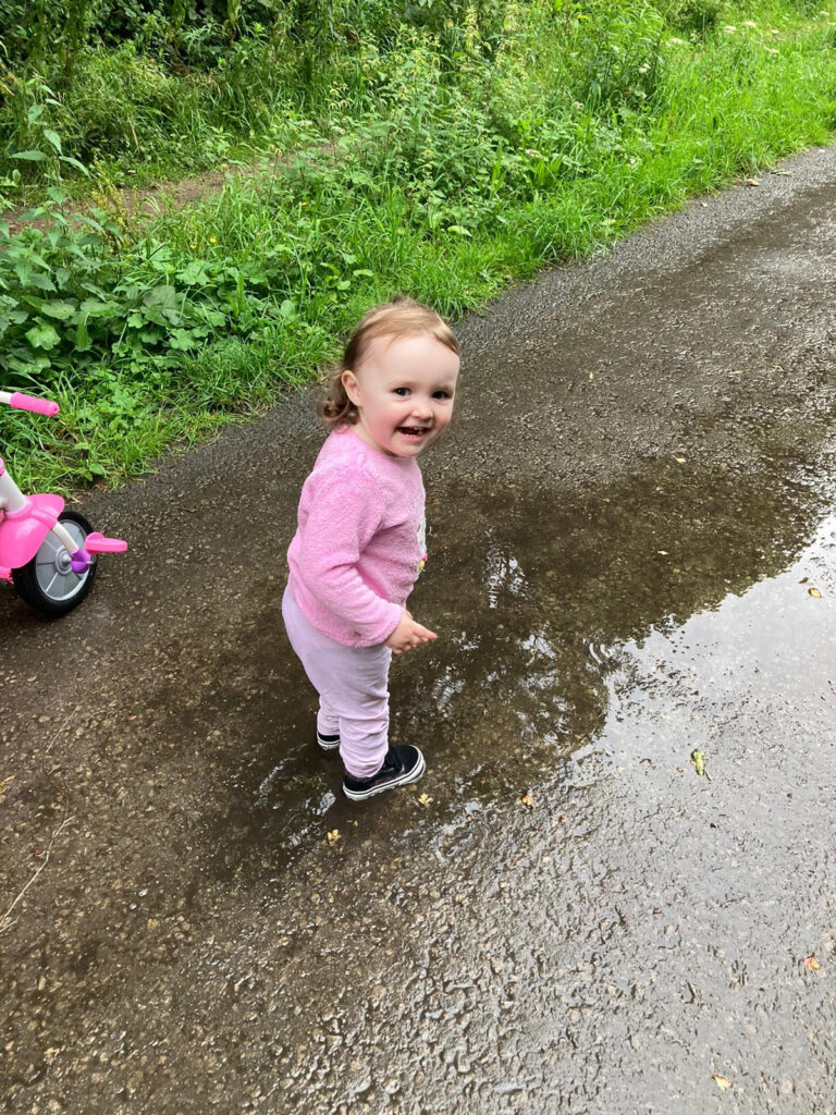 A toddler dressed in pink is splashing in a puddle and smiling at the camera.