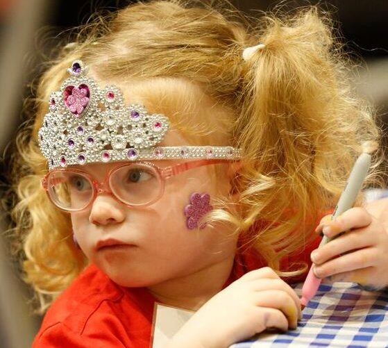 A small child is wearing a crown and pink glasses and drawing.