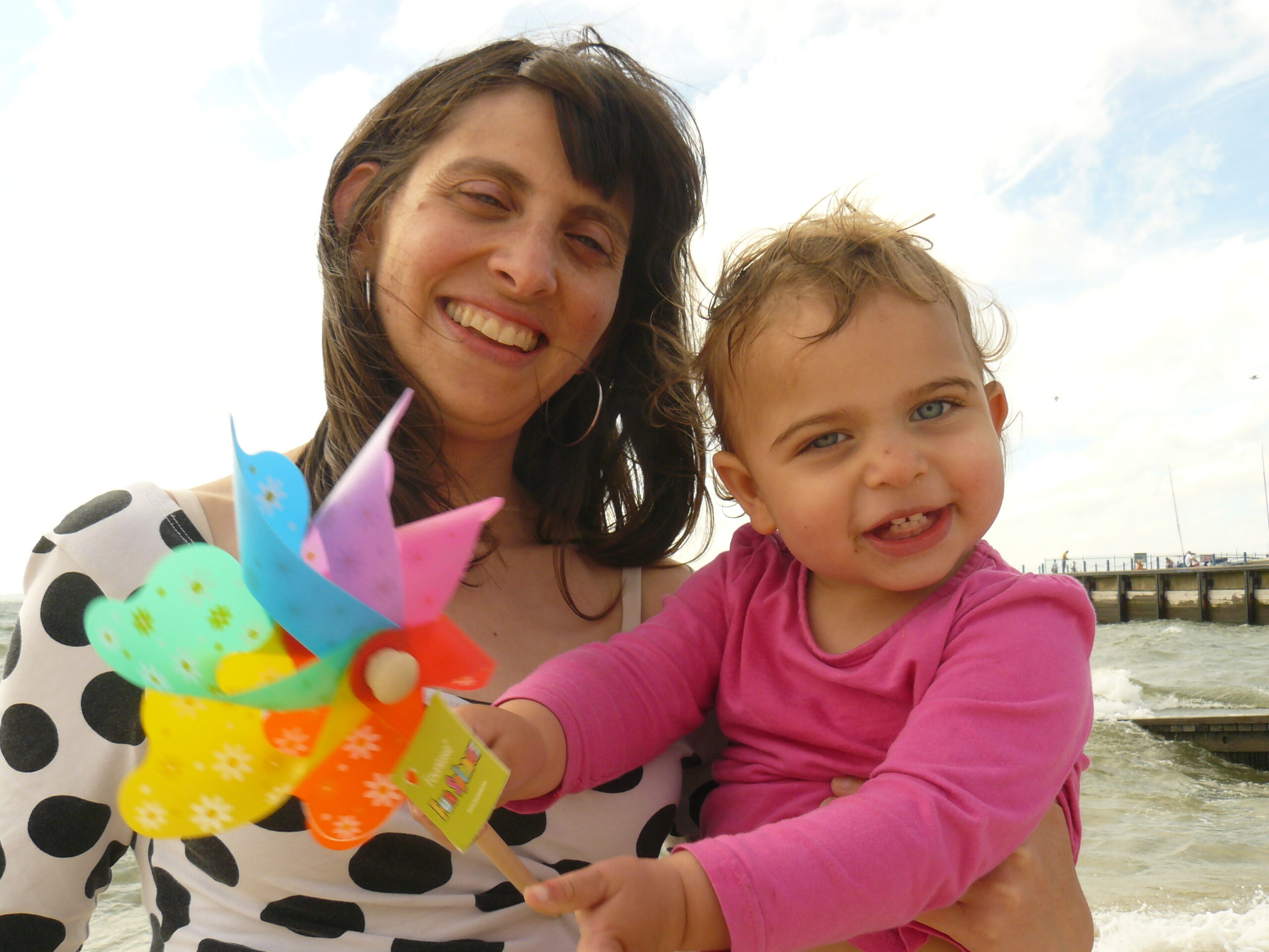 Julia and her young daughter smile for the camera. They are at the seaside and the child is holding a toy windmill.