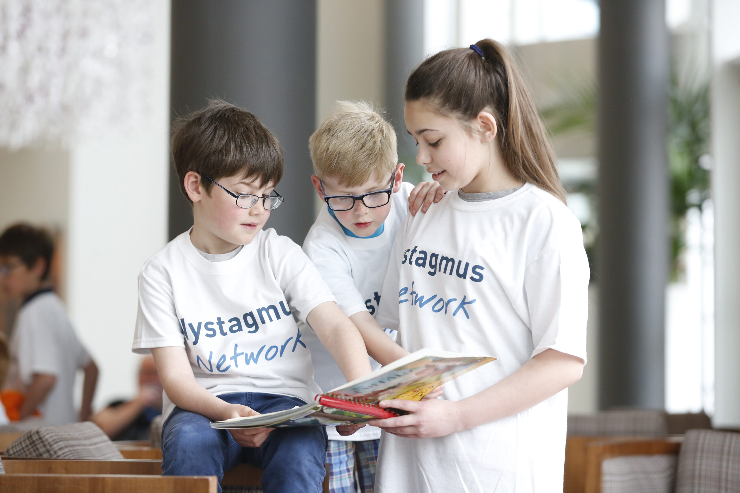 3 children reading a book together. They are all wearing Nystagmus Network T-shirts.