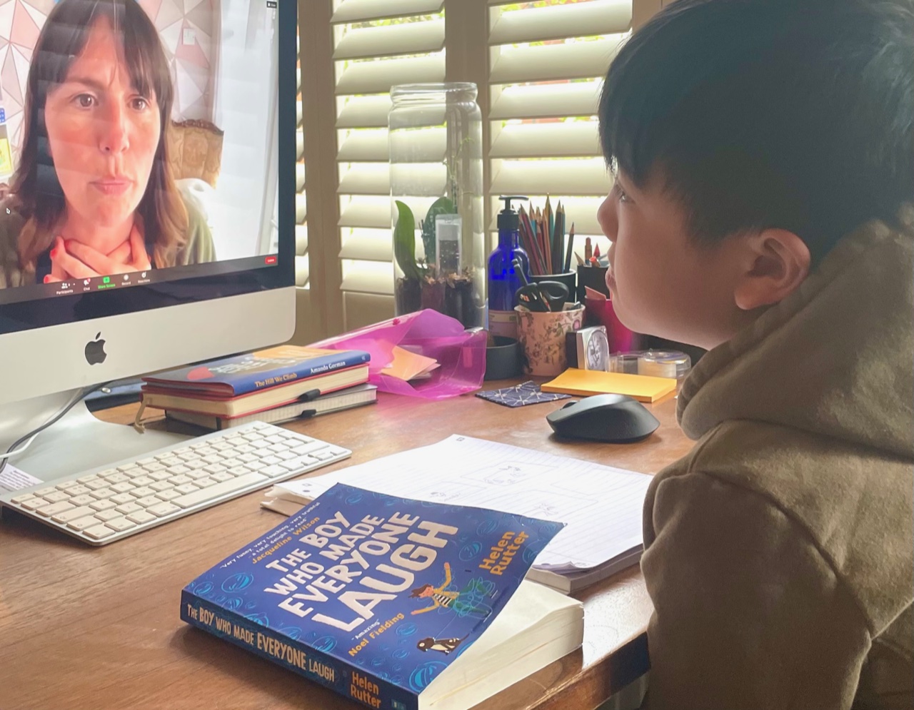 A child takes part in an online writing workshop with author, Helen Rutter. On his desk is Helen's book 'The Boy Who Made Everyone Laugh'.