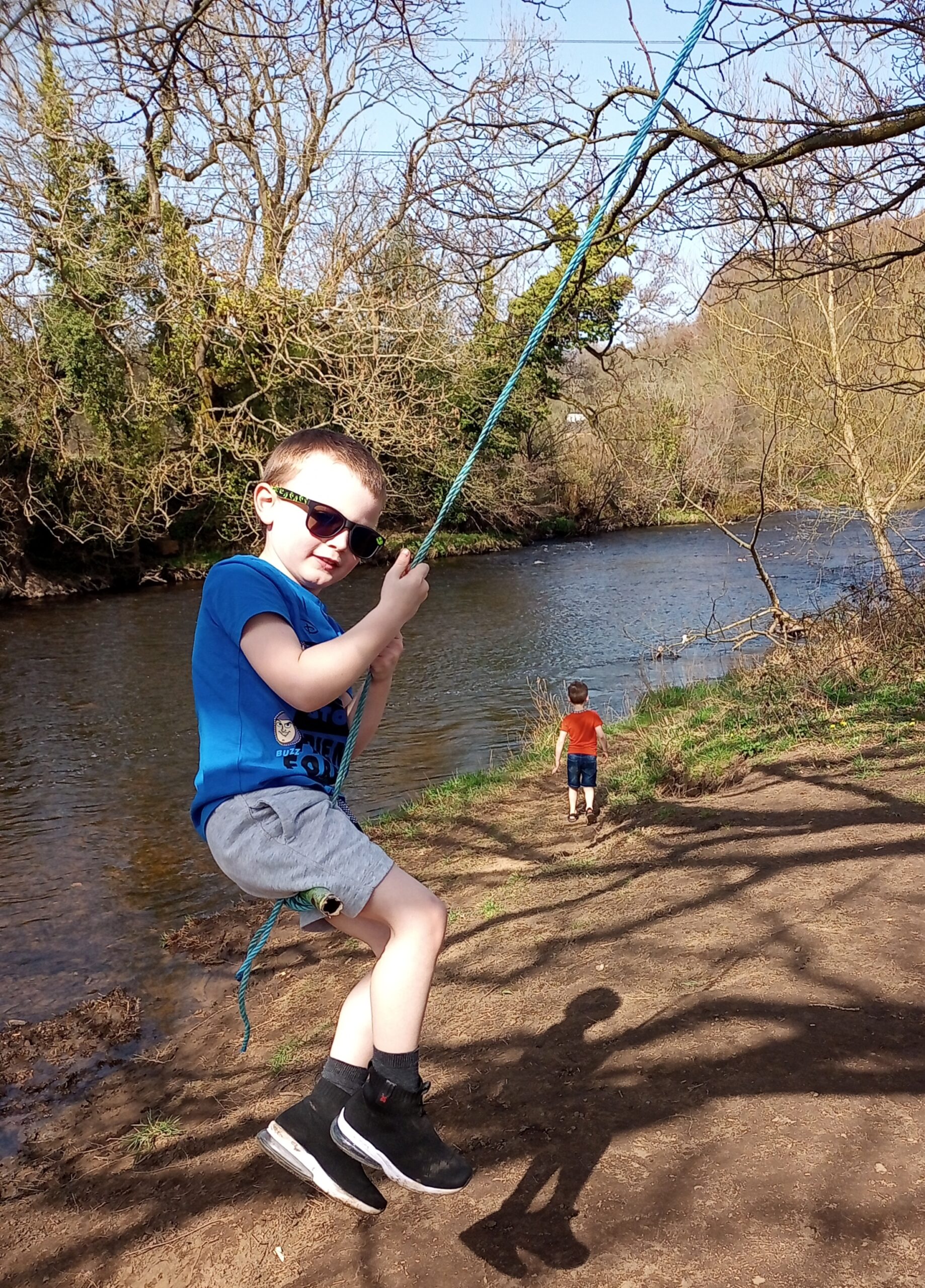 Grayson enjoys a rope swing by a lake. He is wearing dark glasses.