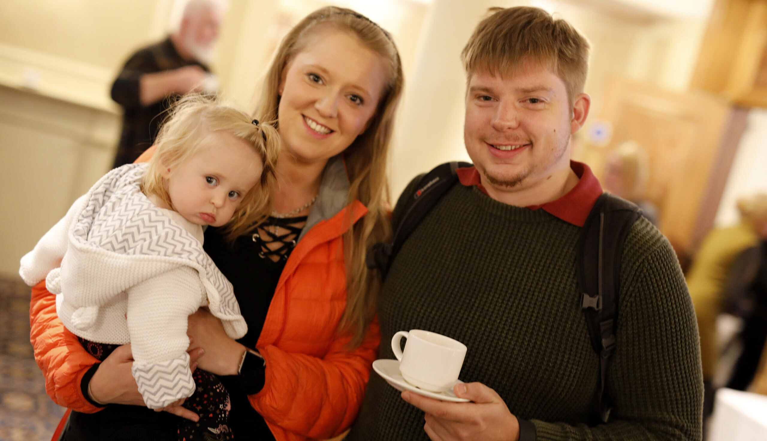 A family smile for the camera. A mother is holding a young child in her arms and the father is holding a cup of tea.