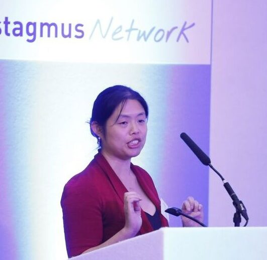 Helena present from a lectern. The Nystagmus Network logo appears on the screen behind her.