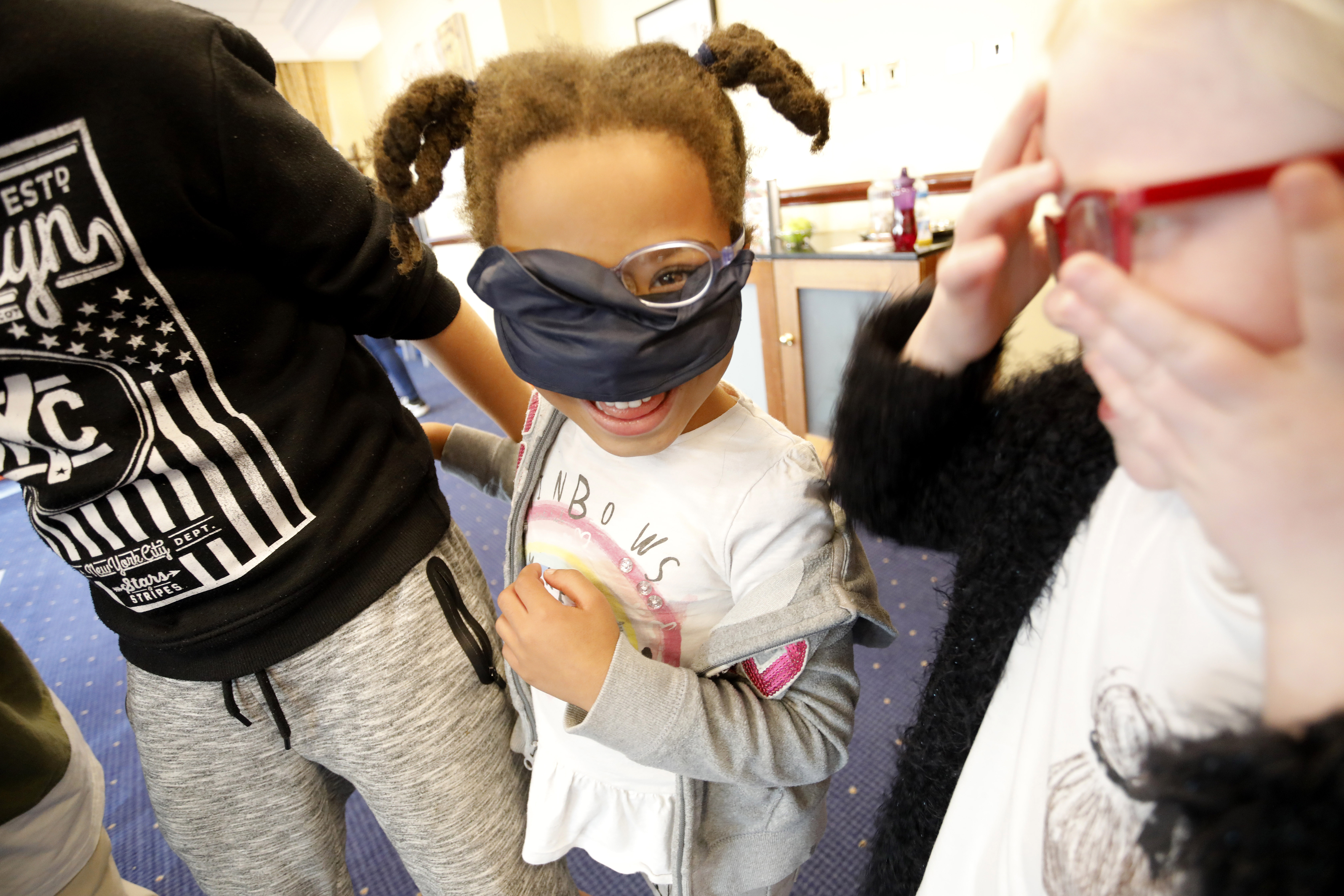 A child playing with her friends. She is wearing a blindfold over her glasses, but looking out over the top with one eye. She is laughing.