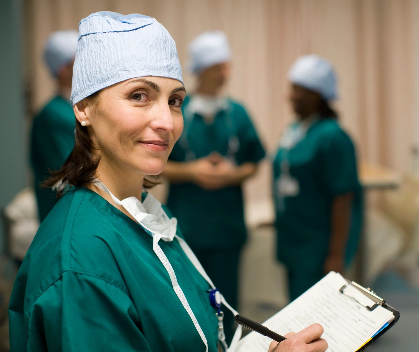 A doctor wearing scrubs is writing on a clipboard. She is smiling at the camera.