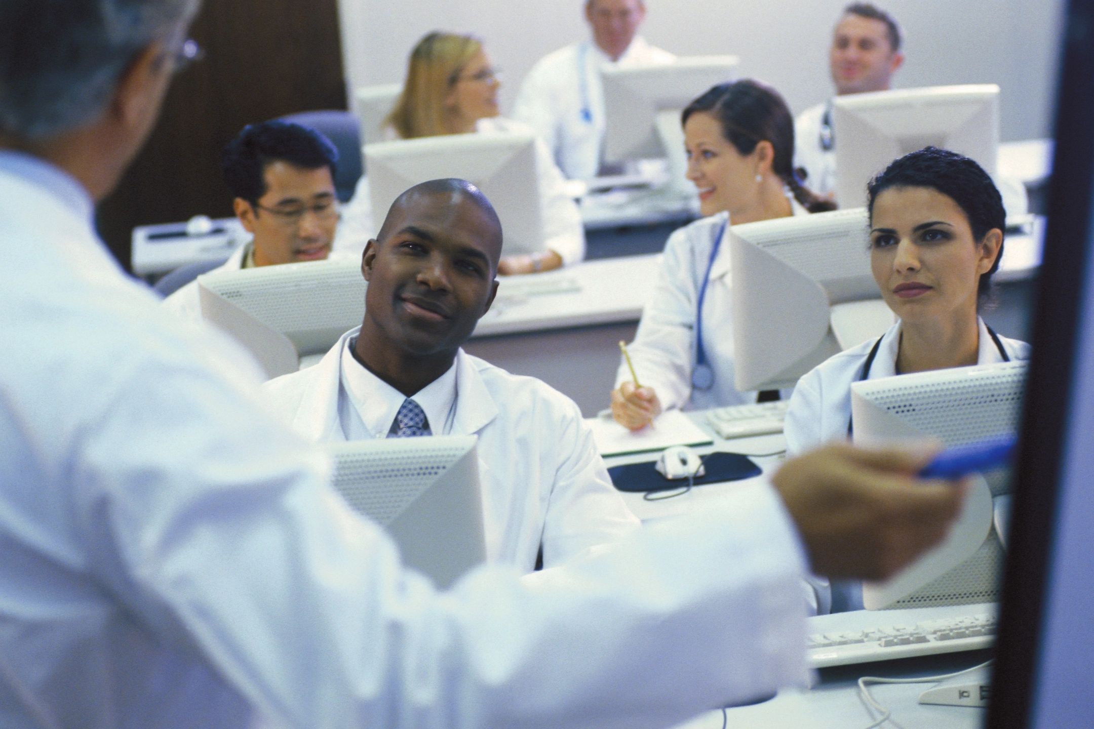 A group of people wearing white coats sit in rows behind laptops with a lecturer pointing at a screen.