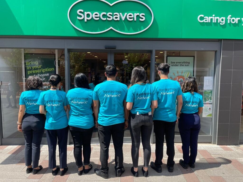 A group of people wearing Nystagmus Network T-shirts standing with their backs to the camera outside a branch of Specsavers.