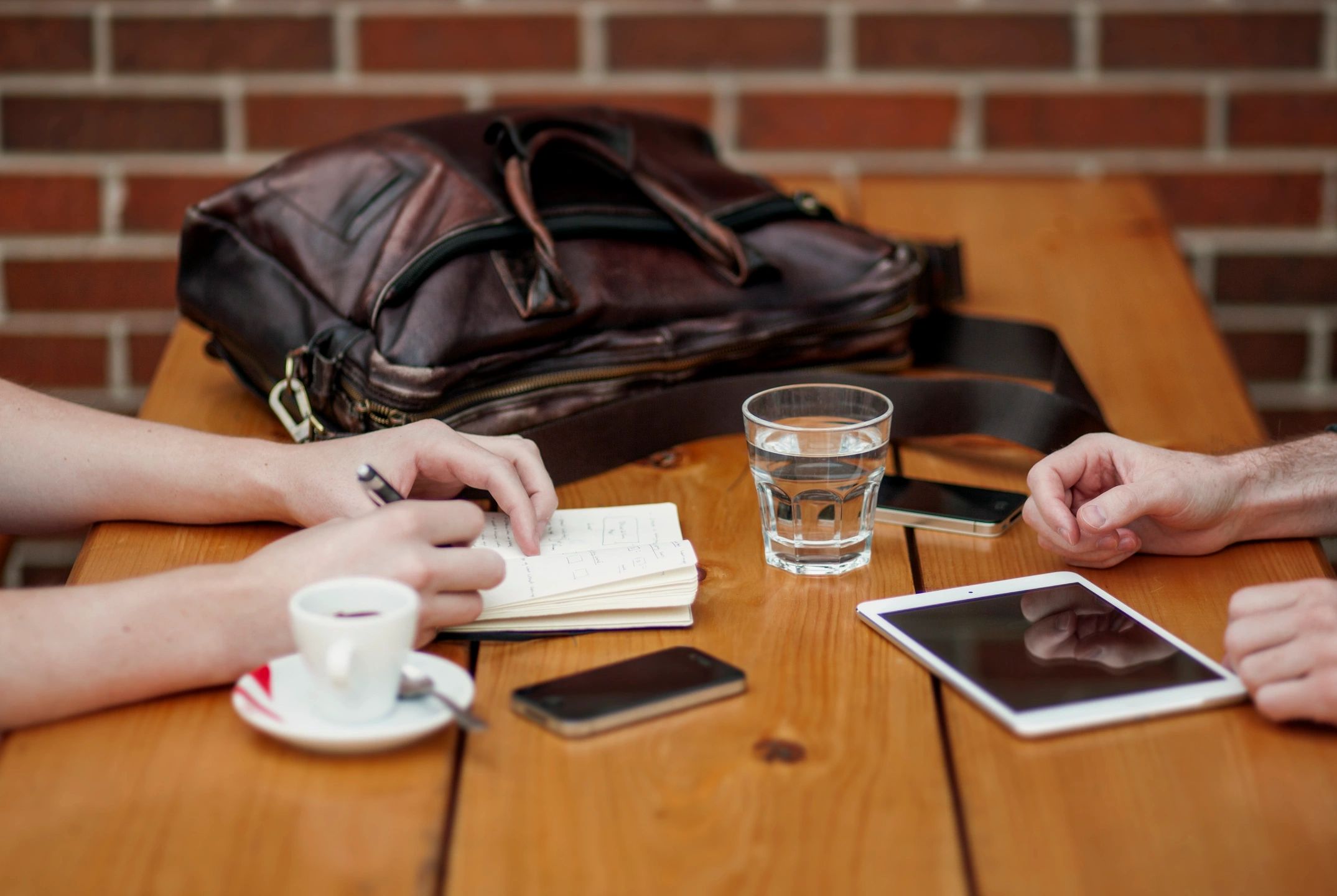 Two people sitting at a meeting table. We see only their hands, a notepad, an electronic device, a coffee and a glass of water.