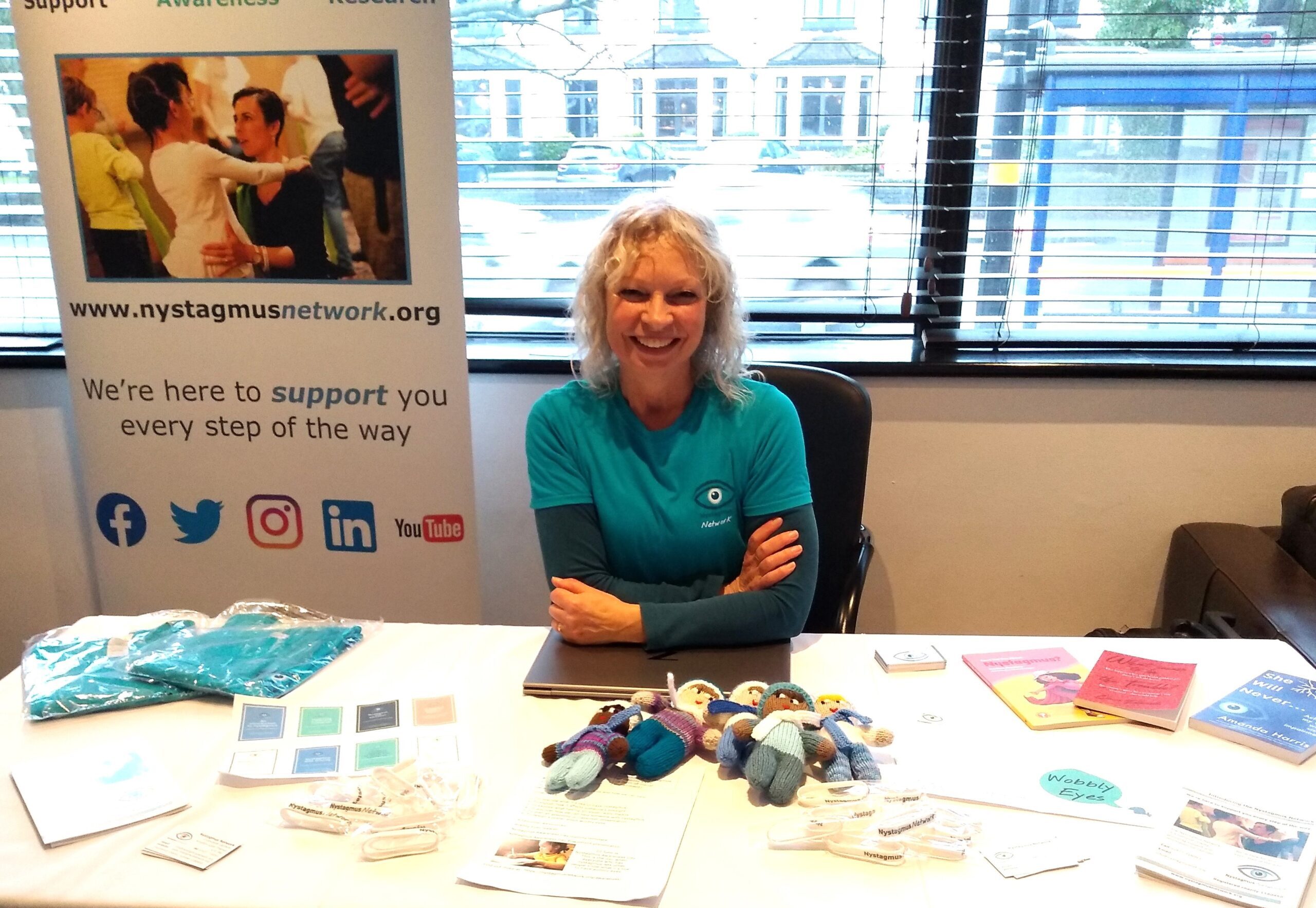 Sue wears a Nystagmus Network T-shirt and sits at her exhibition table.
