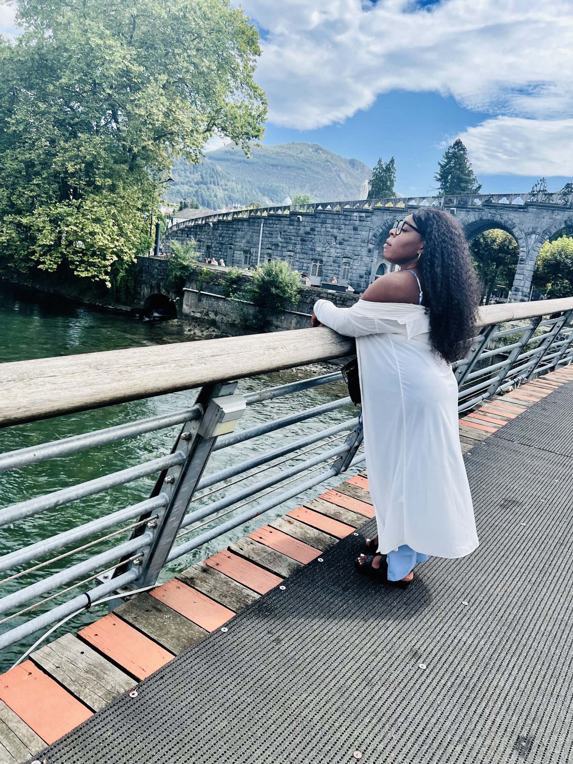 A picture of Sherifa wearing a white long sleeve shirt, baby blue trousers, black large framed glasses, black curly hair and black sandals. Standing on a bridge looking towards a flowing river.