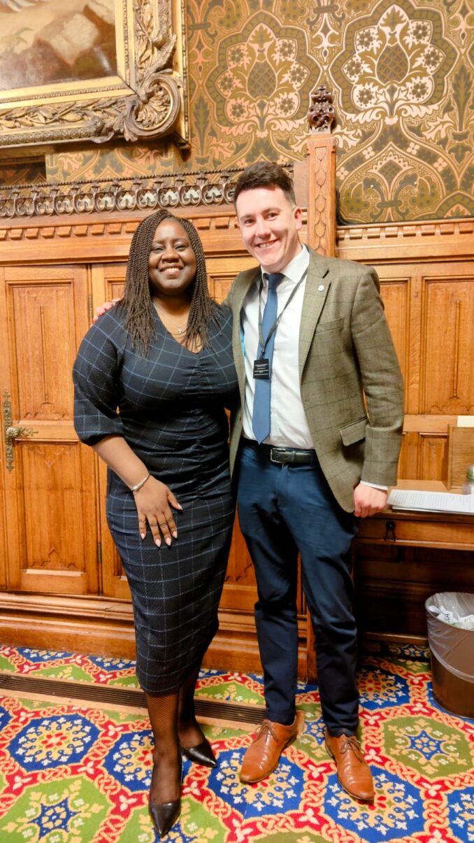 Marsha and George stand together in a parliamentary council chamber.