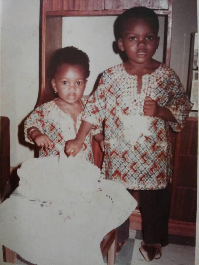 Two young children in party clothes stand beside a frosted cake.