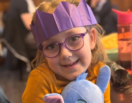 A young child wearing a purple paper crown and glasses is cuddling a blue toy and smiling for the camera.