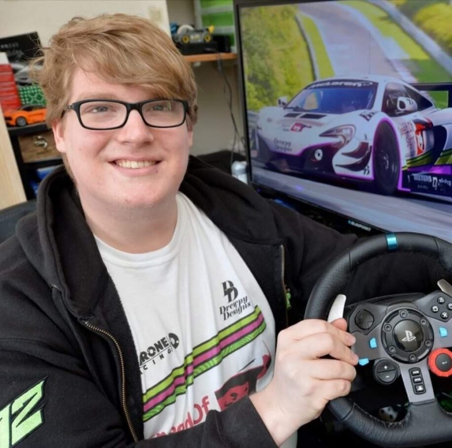 Drew is wearing glasses and is smiling for the camera holding a steering wheel. In the background there is a rally car.