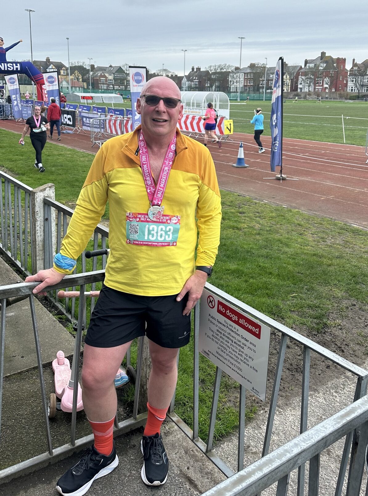 Glen proudly standing in front of a running track wearing a medal. He is wearing sunglasses and a bright yellow jacket.