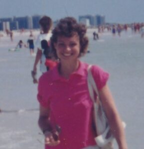 Sue weas a pink polo shirt and carries a beach bag, sunglasses and her shoes. She is on a beach.