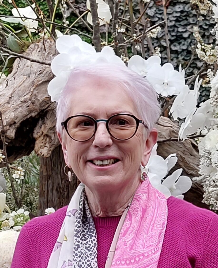 Jan is smiling for the camera, wearing glasses, a pink blouse and a patterned scarf