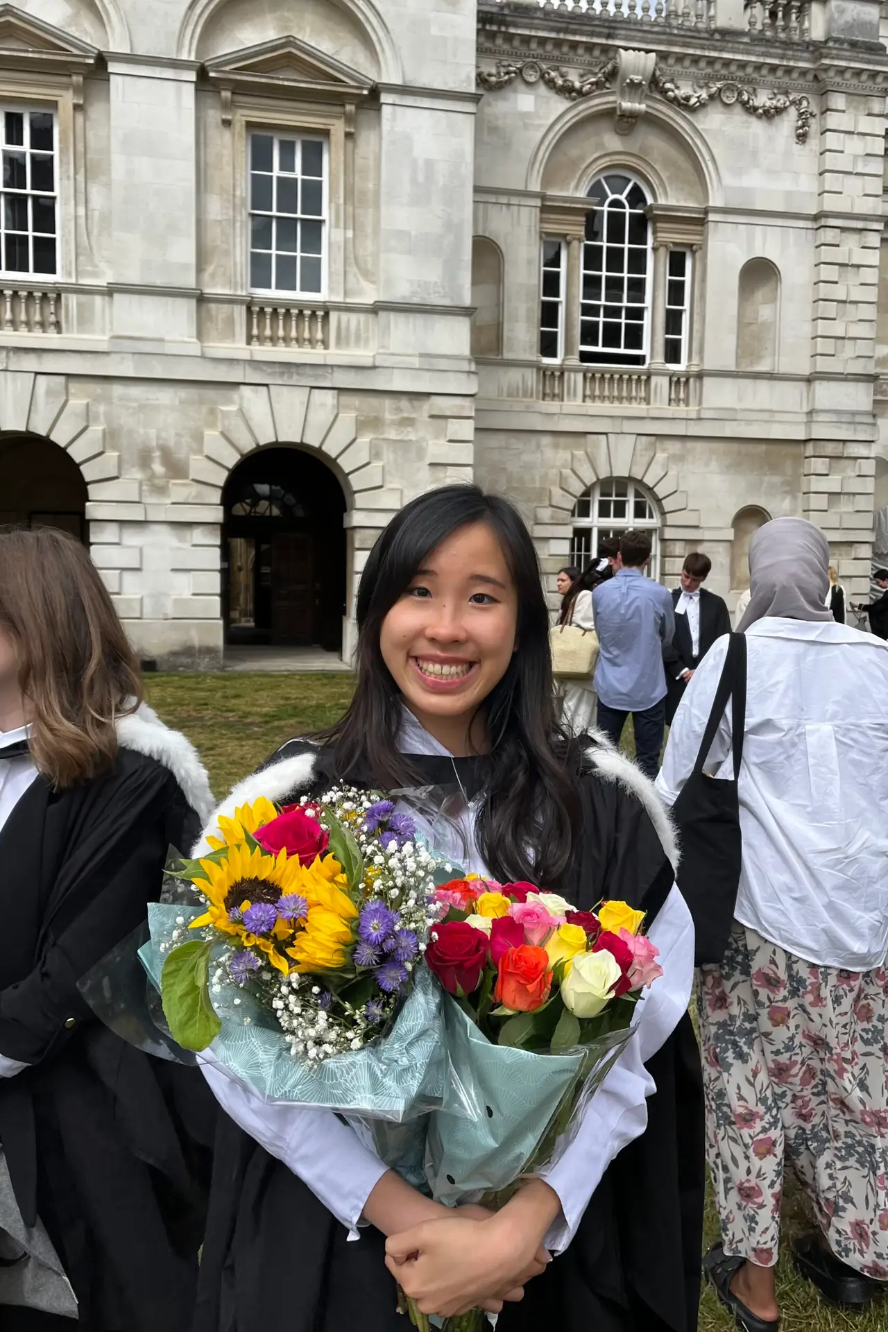 Justine wears cap and gown and holds a bouquet of flowers. She stands in front of a Cambridge University building and smiles for the camera.