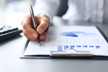 A hand olding a pen, hovering over a graph on a page on a clipboard.