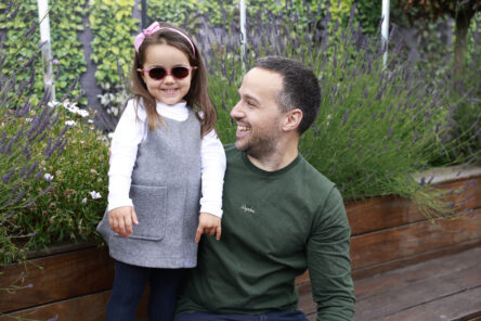 A young child wearing dark glasses is standing in a garden beside her father.
